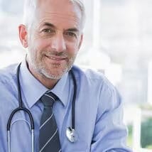 Smiling male doctor with gray hair wearing a light blue shirt, striped tie, and stethoscope around his neck, looking at the camera in a bright clinical setting.