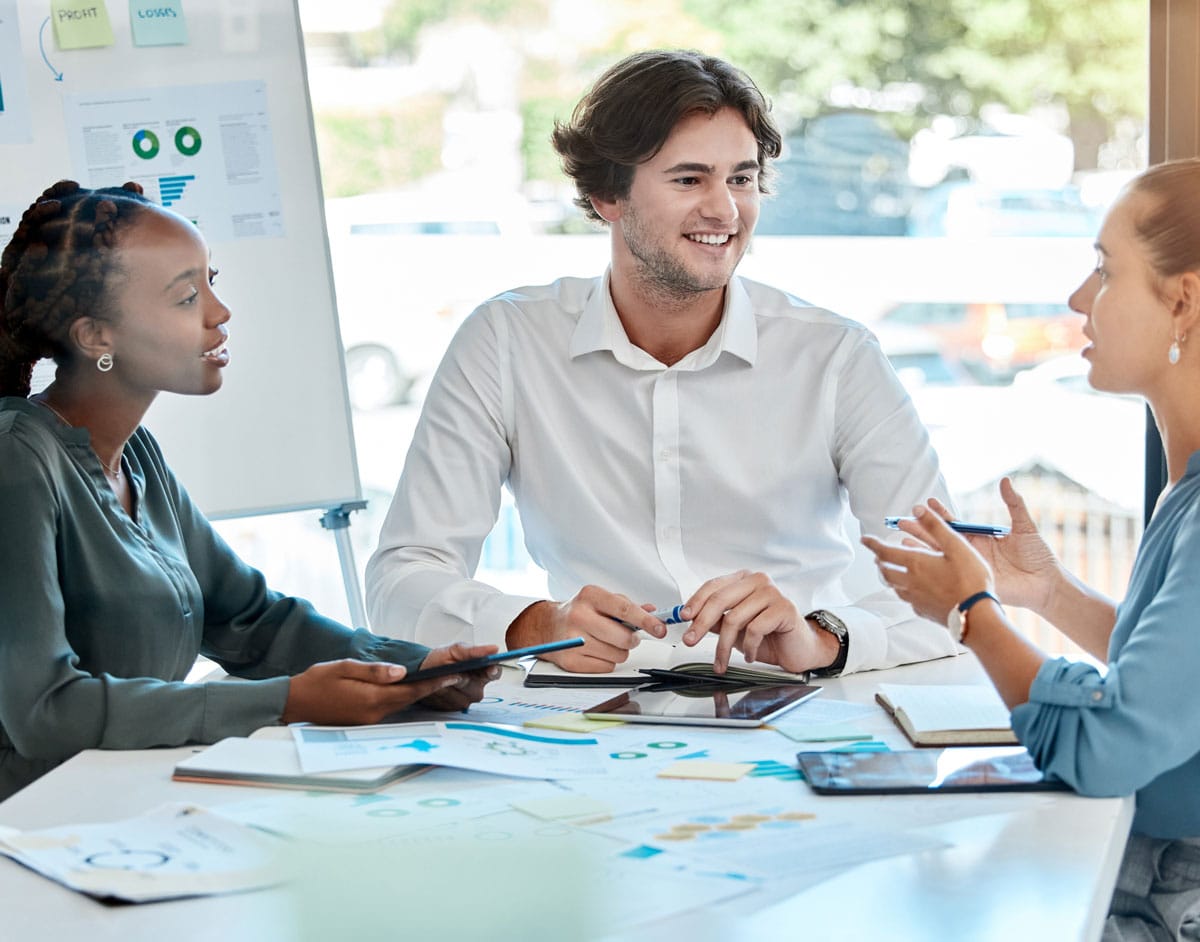 Two women and a man talking over a table with charts on paper and tablets