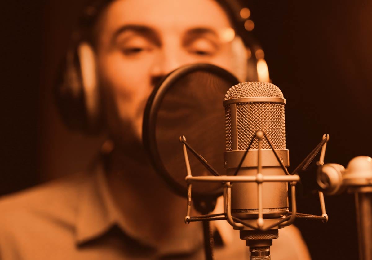 Close-up of a studio condenser microphone with a pop filter in warm lighting, with a male vocalist wearing headphones blurred in the background. A hero image for home vocal recording and audio tracking.