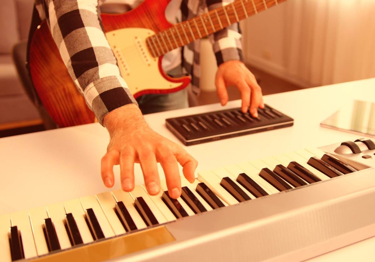 Close-up of a musician's hands playing a white MIDI keyboard controller on a desk while wearing an electric guitar. A hero image showcasing essential gear for home music production.