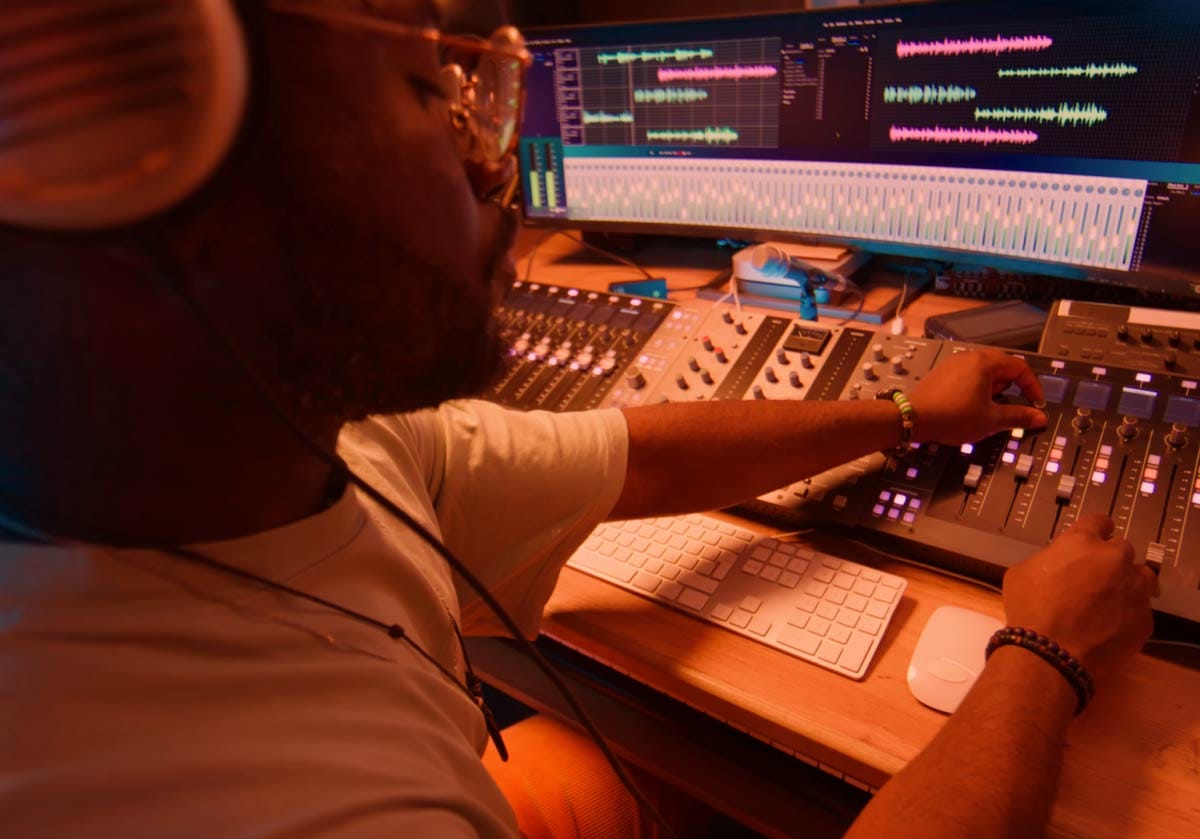 Side profile of a music producer wearing headphones and glasses, adjusting a mixing console in a studio with dual monitors displaying audio waveforms. A hero image for modern music production and beatmaking.