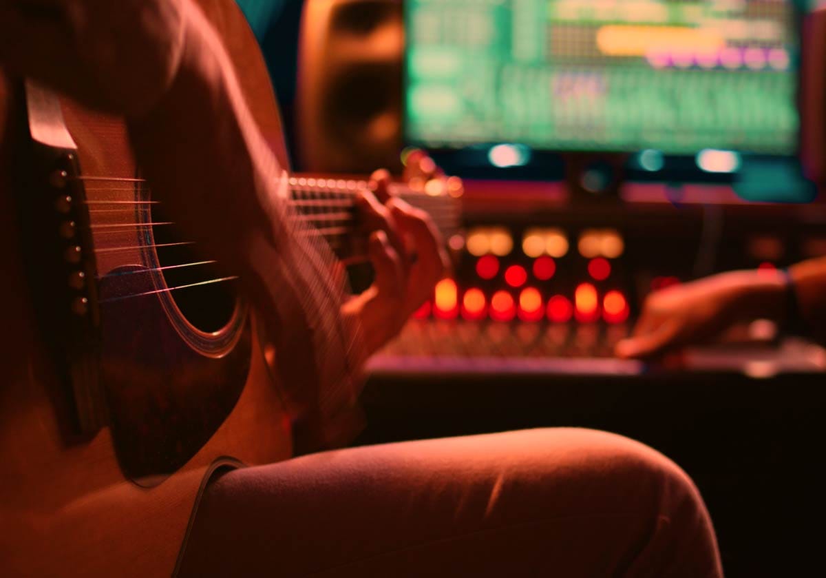 Close-up of a musician playing an acoustic guitar in a home studio, with a glowing digital audio workstation (DAW) mixer in the background. A hero image representing songwriting and composition skills for modern music creators.