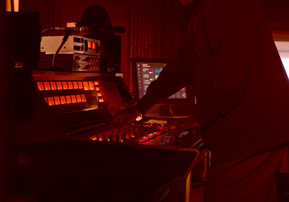 A music producer adjusting a classic analog mixing console inside a dark, moody home studio illuminated by deep red and orange ambient accent lighting. A hero image representing affordable home studio setups.
