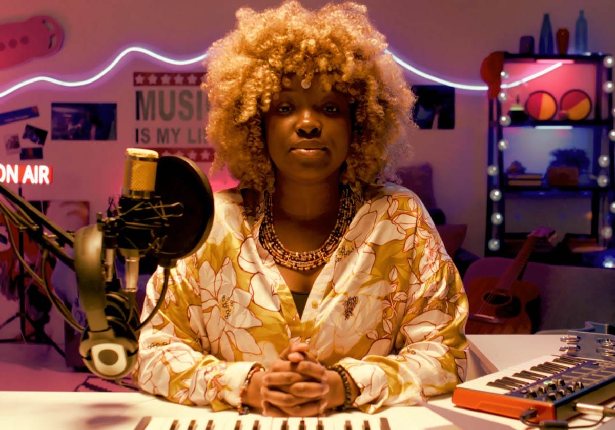 A female music creator with curly blonde hair sitting at a studio desk behind a condenser microphone and MIDI keyboard controller, with colorful ambient studio lighting in the background.