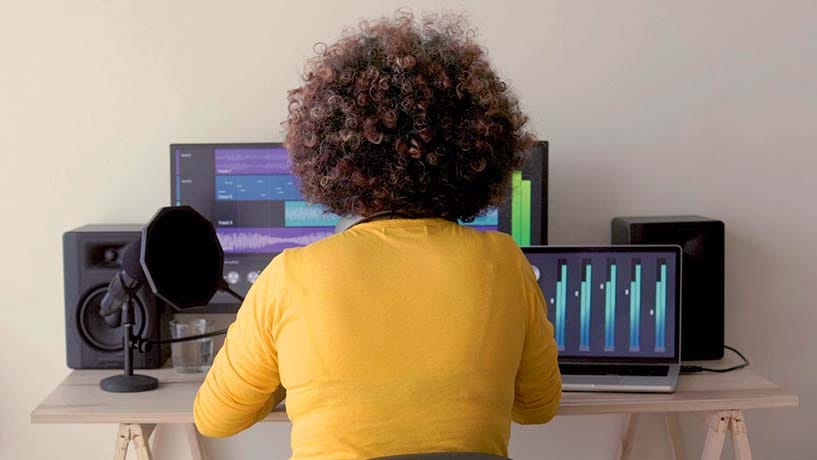 A music producer working at a dual‑screen studio setup with speakers and a microphone, illustrating the course’s focus on musicians who want deeper control over MIDI realism and professional workflow.