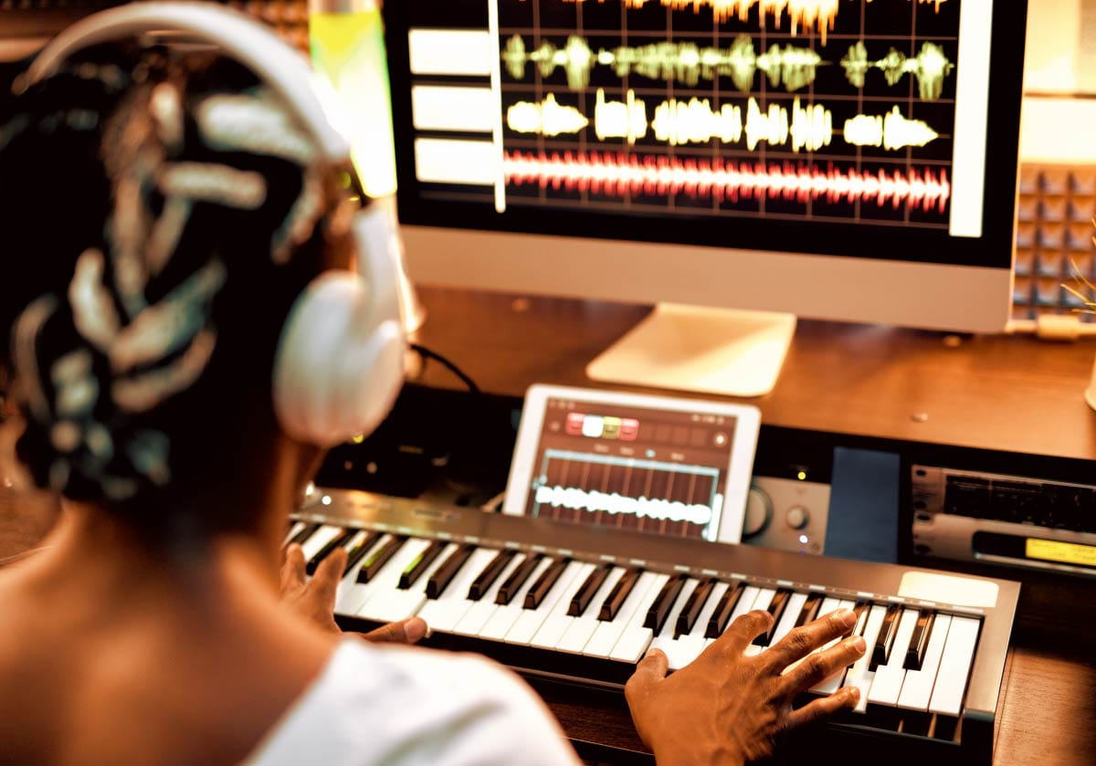 Over-the-shoulder shot of a music producer wearing white headphones at a desk, playing a MIDI keyboard controller while looking at an audio waveform timeline on a desktop monitor.