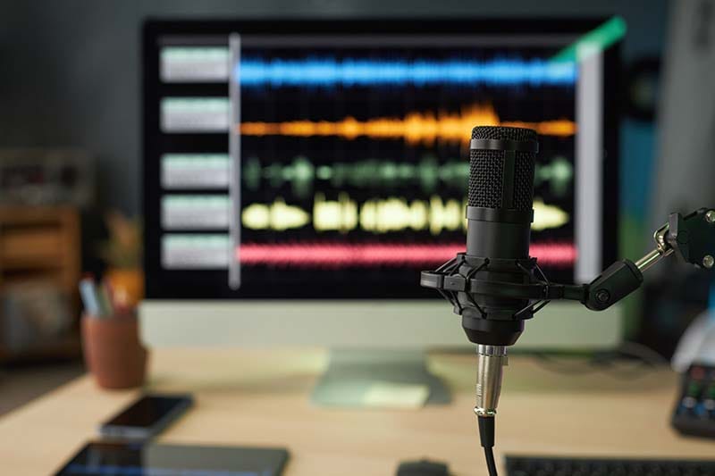A desktop studio workspace featuring a black condenser microphone on a boom arm in sharp focus, with a computer monitor displaying colorful multi-track digital audio waveforms in a DAW blurred in the background.