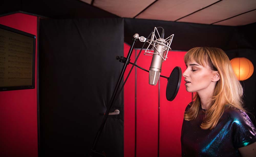 Side profile of a female vocalist singing into a large-diaphragm condenser microphone with a pop filter inside a home recording studio equipped with red and black sound absorption wall panels.