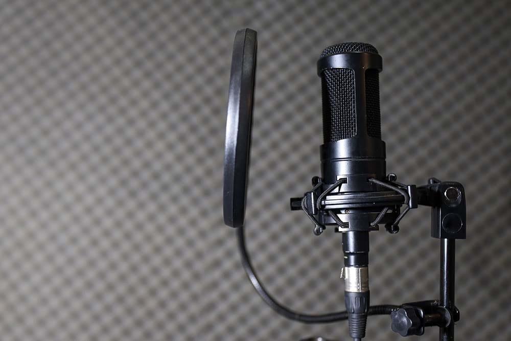 A professional black condenser microphone securely mounted in an elastic shock mount with a round pop filter positioned in front of a grey acoustic foam tiled wall.