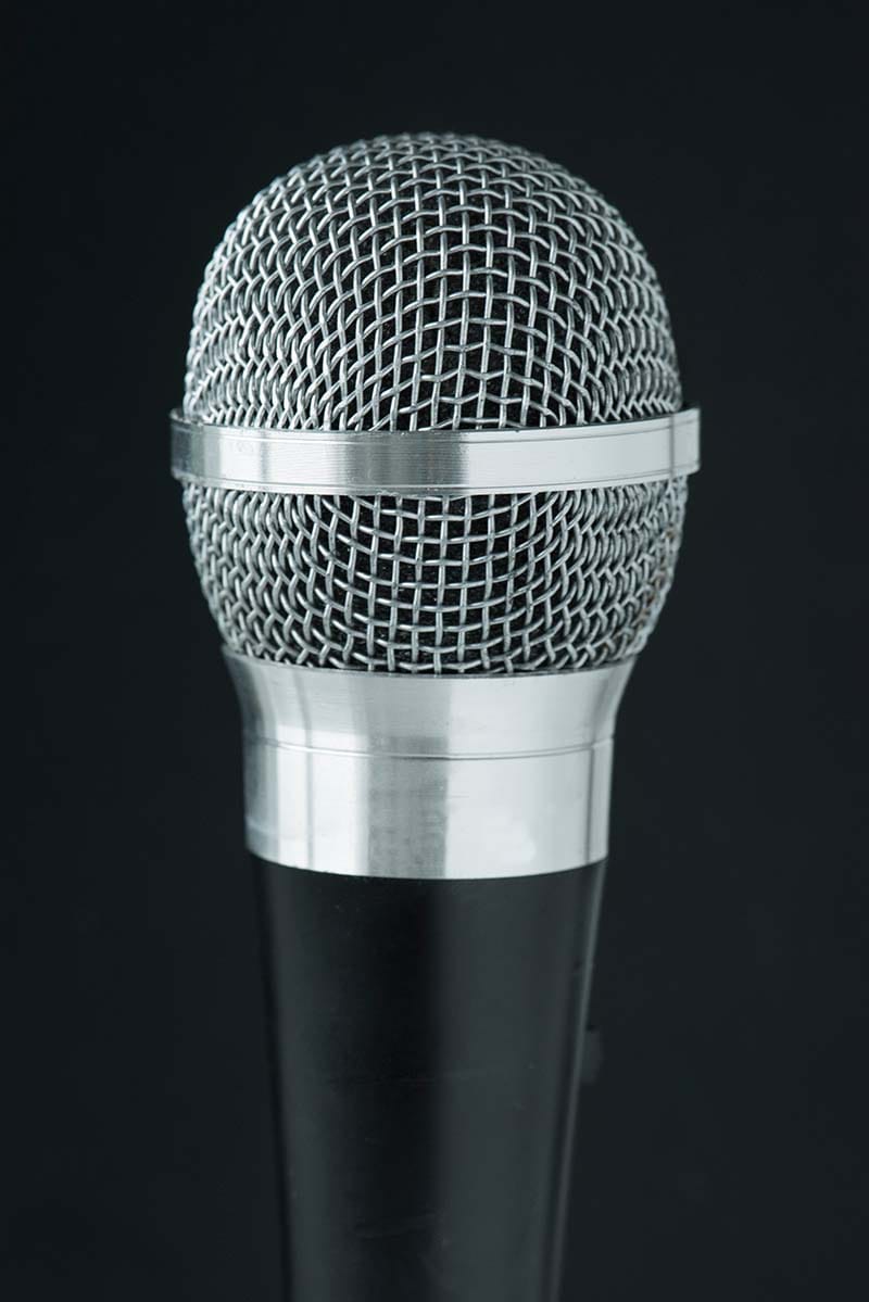 Close-up shot of a classic handheld dynamic microphone featuring a textured silver steel mesh grille and a matte black handle against a dark background.