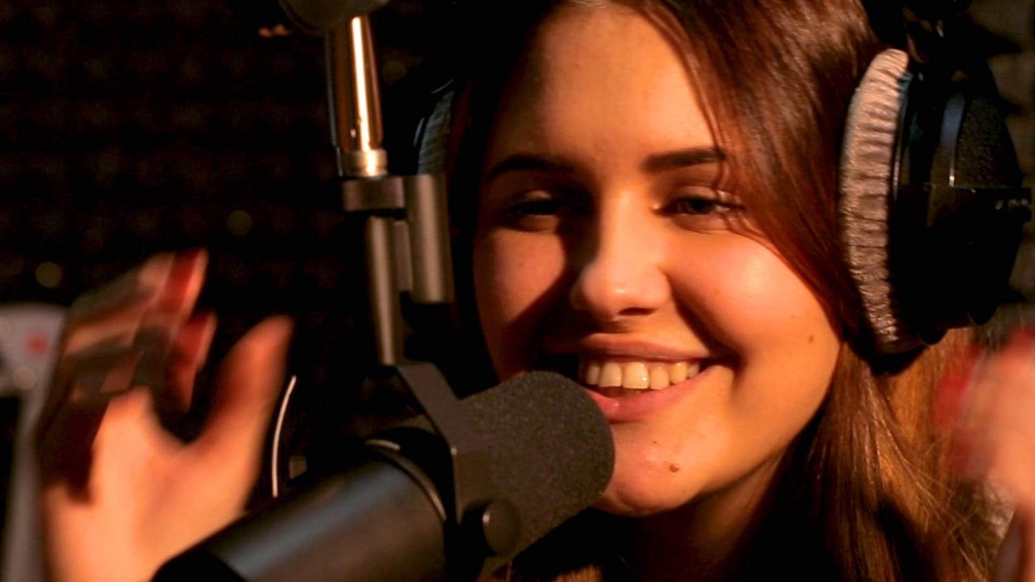 Close-up of a smiling, confident female vocalist wearing studio headphones, singing into a Shure SM7B dynamic microphone in a dimly lit home recording booth.