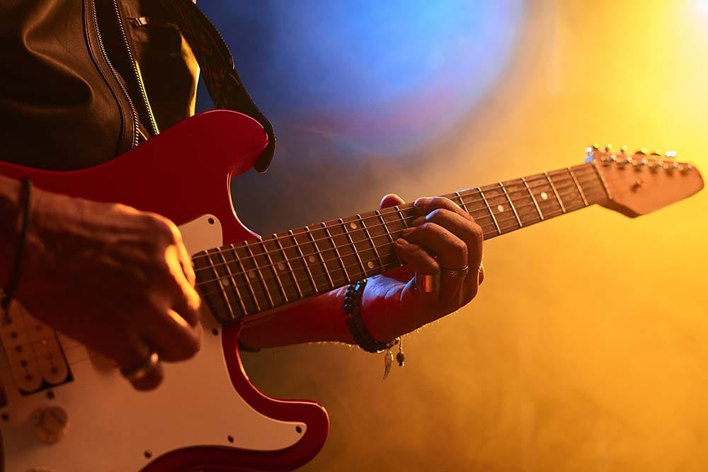 A close-up view of a guitarist's hands playing chords on a red electric guitar on a dark stage illuminated by warm yellow and blue spotlights.