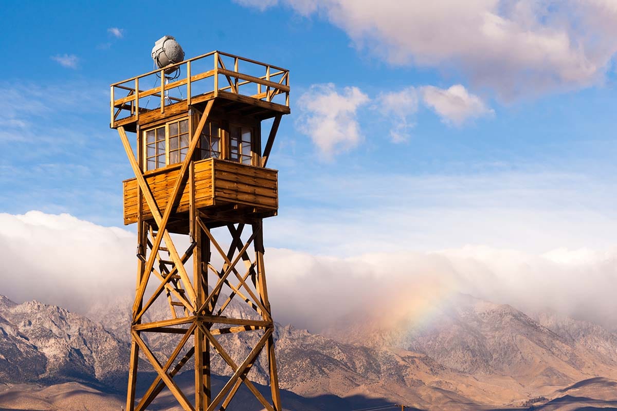 A tall, rustic wooden watchtower stands high under a bright blue sky with passing white clouds. In the background, a faint rainbow stretches over a massive mountain range representing chords that have a lifted feel.