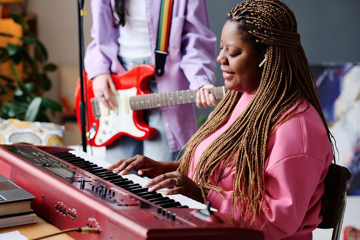 A smiling musician with long braided hair wearing a pink sweatshirt plays a red keyboard in an apartment studio space. In the background, another band member stands and holds a red electric guitar.
