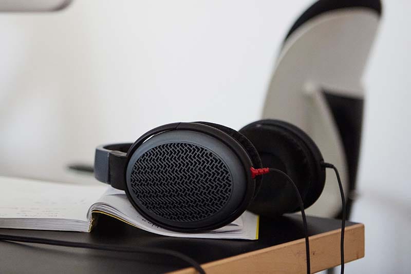 A lifestyle photograph of open-back studio headphones featuring a porous mesh outer grille, resting on an open notebook on a wooden desk.