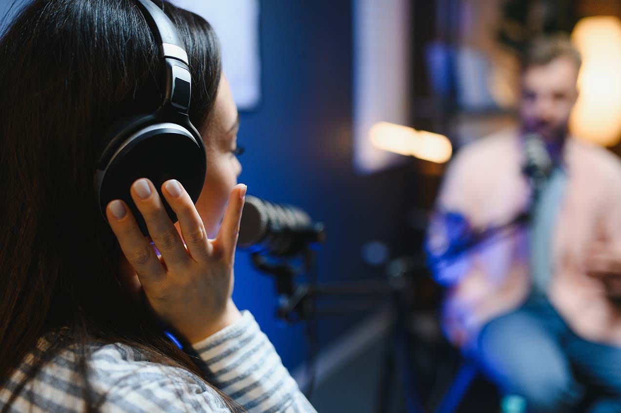 A woman wearing closed-back studio headphones speaking directly into a professional large-diaphragm microphone on a suspension boom arm inside a recording studio.