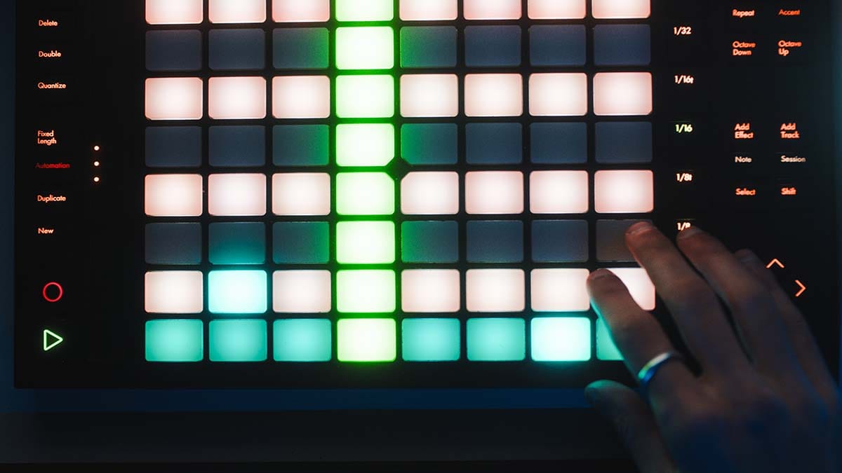Producer's hand playing an illuminated 8x8 grid pad MIDI controller for triggering drum samples and launching clips