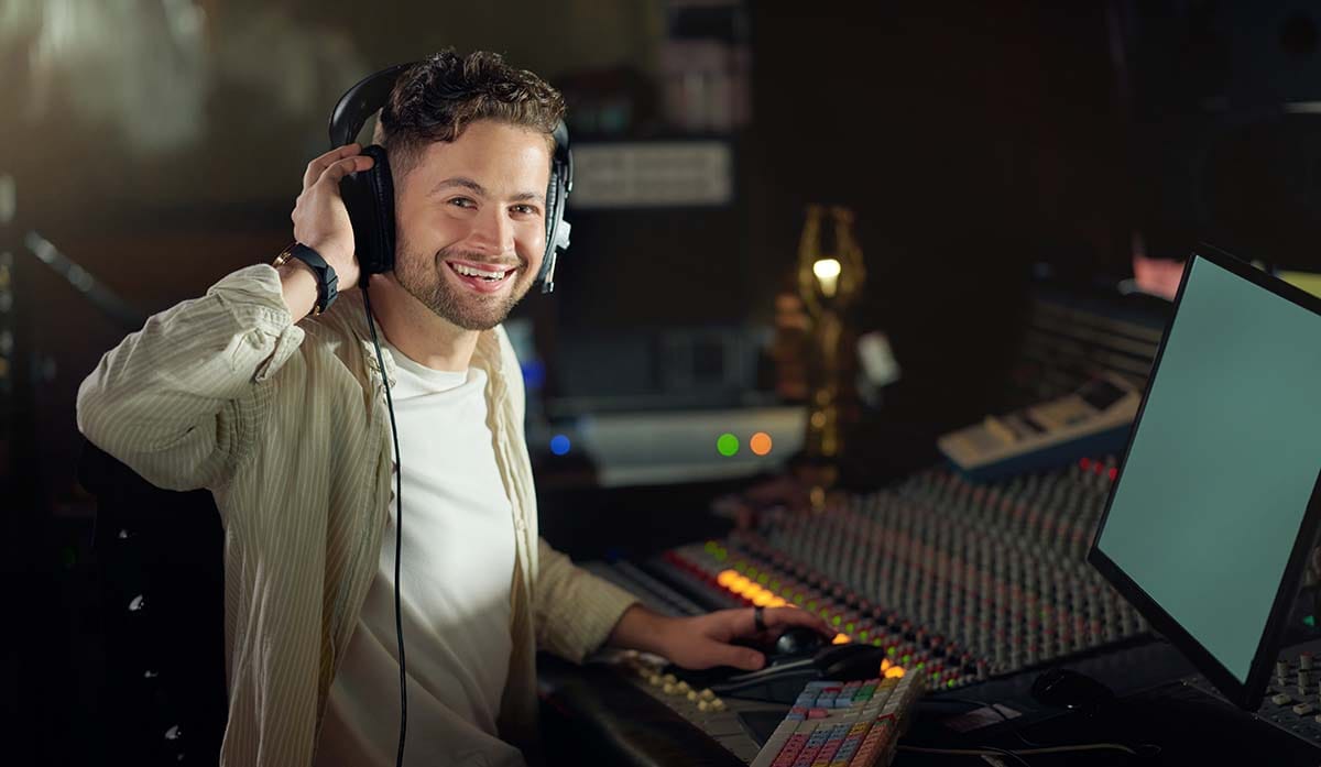 Music producer smiling while wearing studio headphones at a mixing desk in a home recording studio