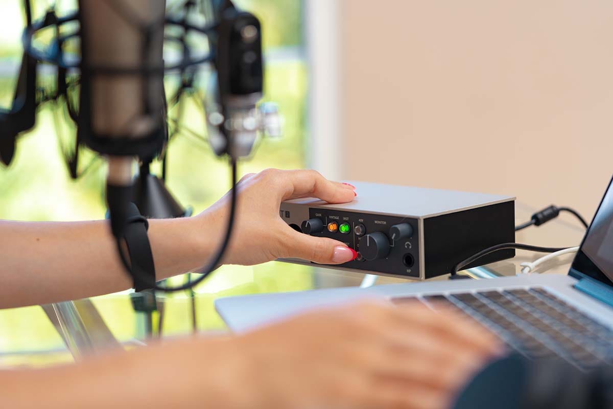Hand adjusting gain controls on a desktop audio interface next to a studio microphone and laptop