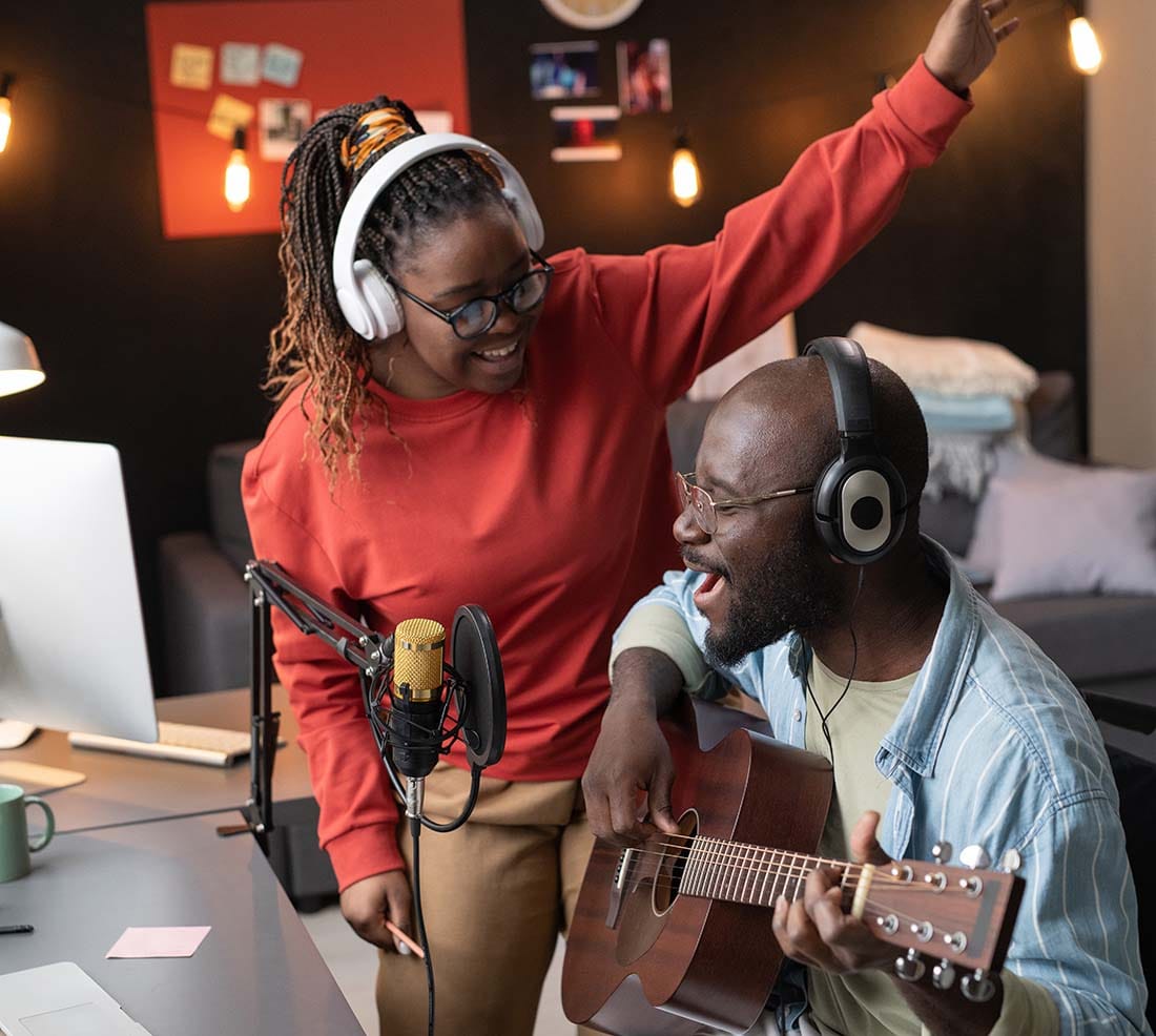 Two music producers laughing and capturing a song idea together in a home recording studio with an acoustic guitar and microphone.