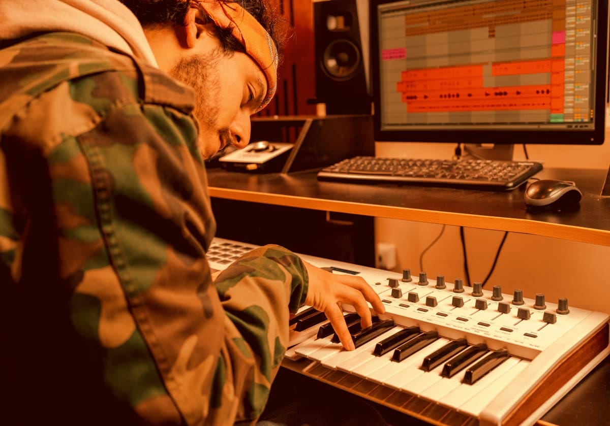 Music producer playing a MIDI controller keyboard at a studio desk while looking at a DAW session on a computer screen.