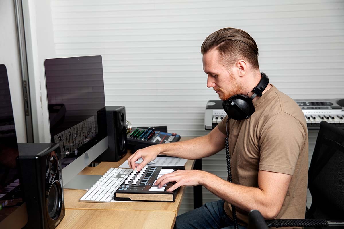 Producer wearing headphones playing a compact MIDI keyboard at a computer desk setup in a home recording studio.