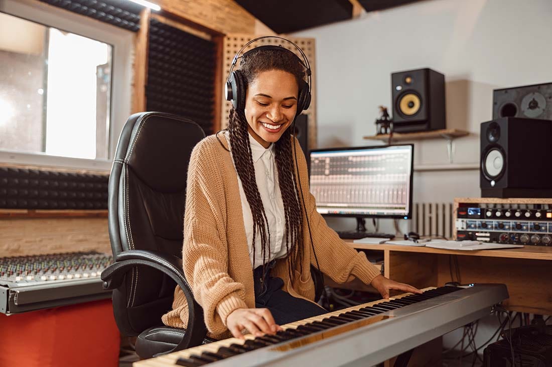 Smiling music producer wearing headphones while playing a keyboard controller in a home studio environment.