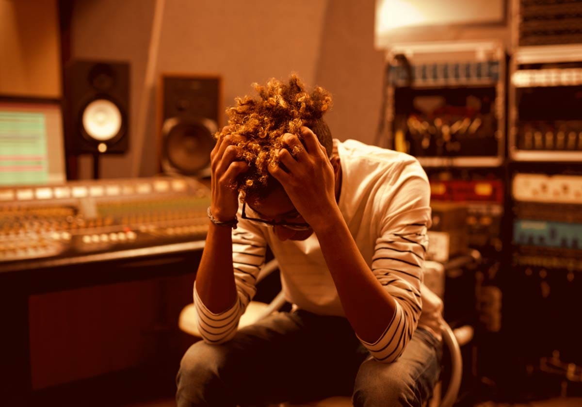 Frustrated music producer sitting in a studio with hands in hair in front of an audio mixing console and studio monitors.