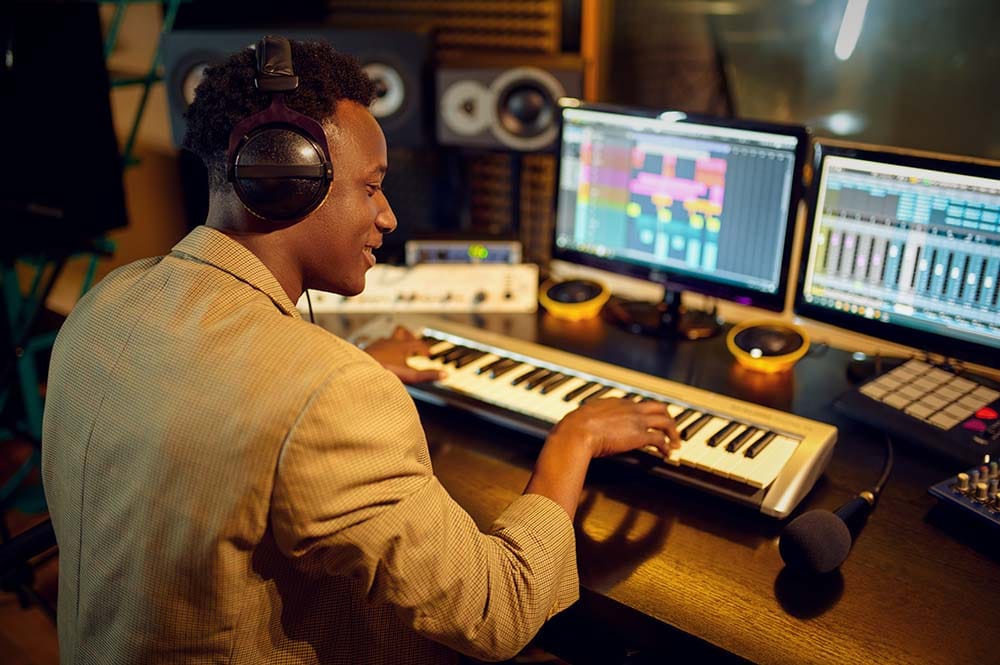 Producer wearing headphones playing a MIDI keyboard at a studio desk with dual computer monitors displaying DAW software.
