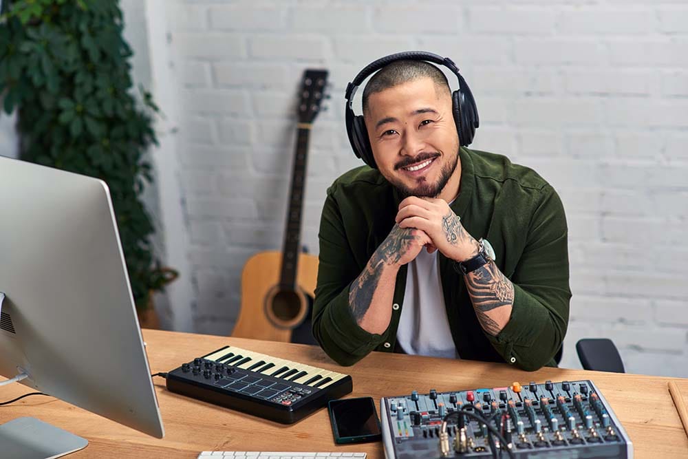 A smiling music producer pictured in their home studio with a MIDI controller, mixing board and guitar.