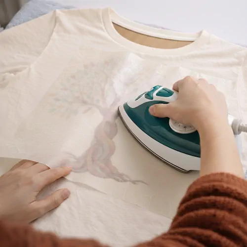 Person ironing a tree design onto a white T-shirt using a transfer sheet and a handheld iron.