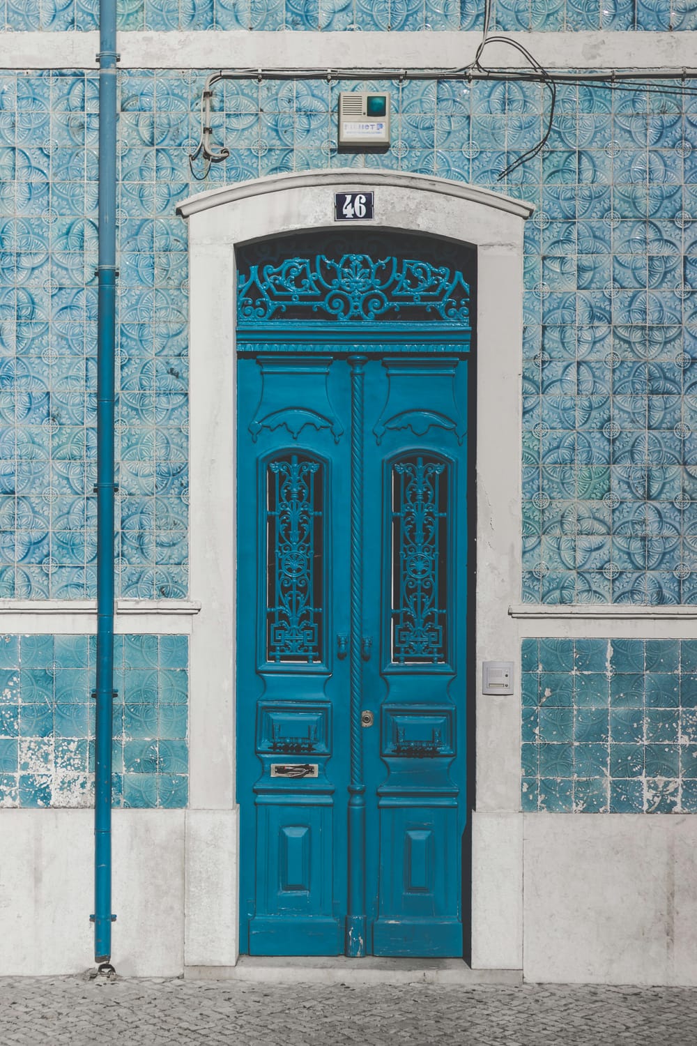 alt="The front doors of a building in Libson, Portugal, featuring a set of double doors with intricate filigrees set against bright blue hand-painted tiles"