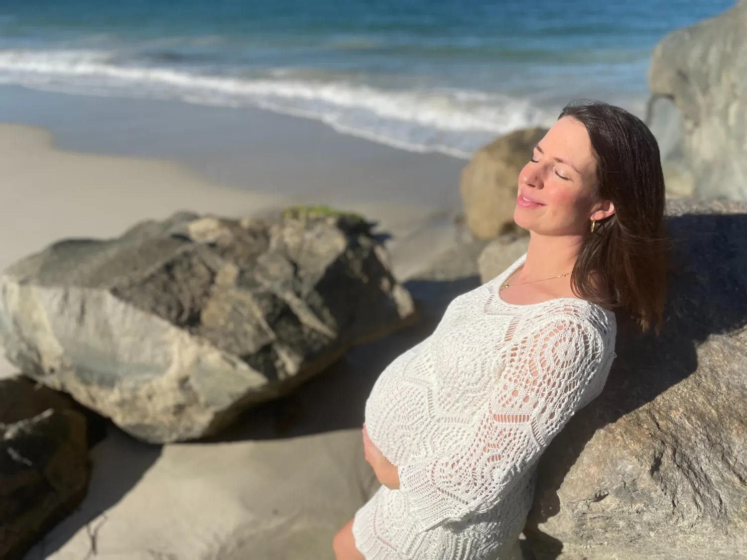 Natalie leaning against beach rocks, pregnant, eyes closed and smiling in the sun