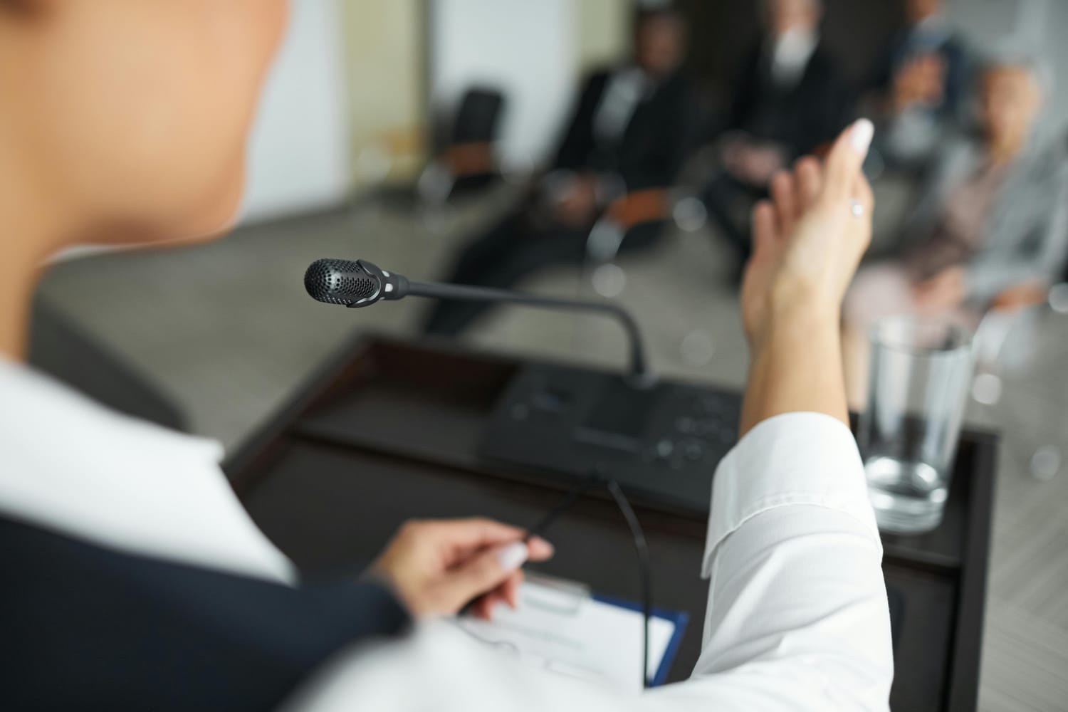An over the shoulder image of a speaker talking into a microphone with a blurred audience in the background. 