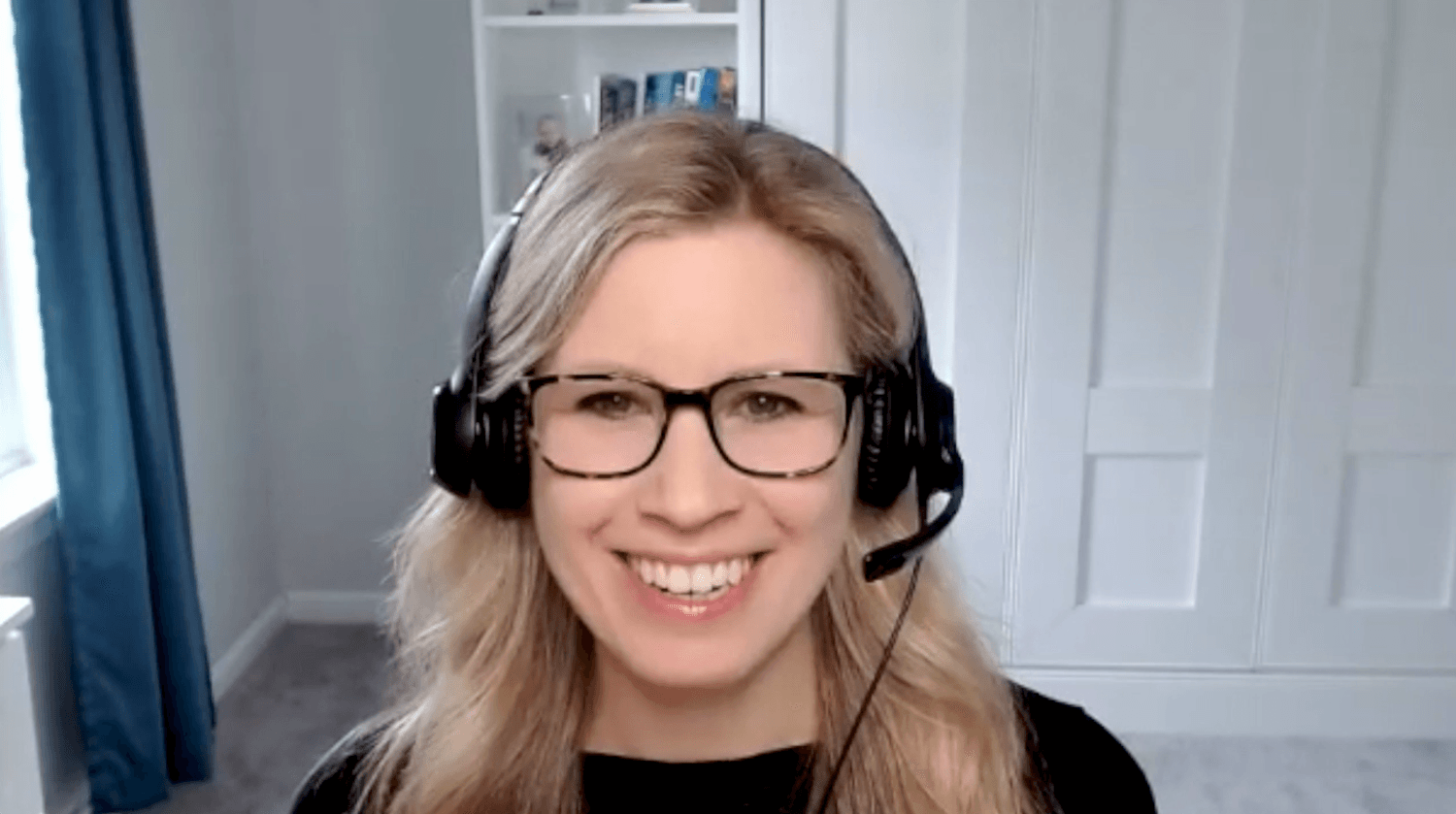 Image of Megan Kohler, blonde hair with black glasses, wearing a black sweater and a headset with a microphone. 