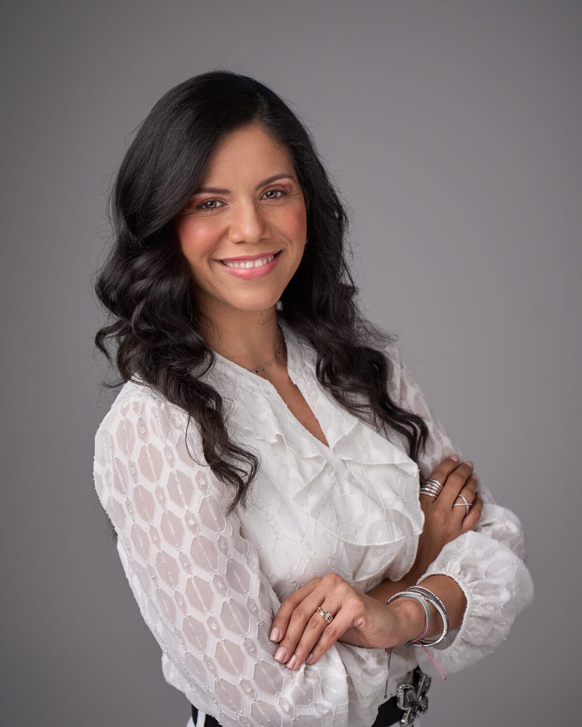Yolanda Rentas sonríe con los brazos cruzados, blusa blanca sobre fondo gris