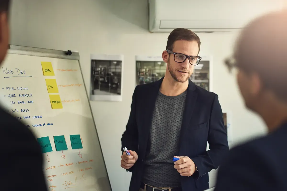 A man in glasses presents a startup pitch deck on web development, with notes on a board.