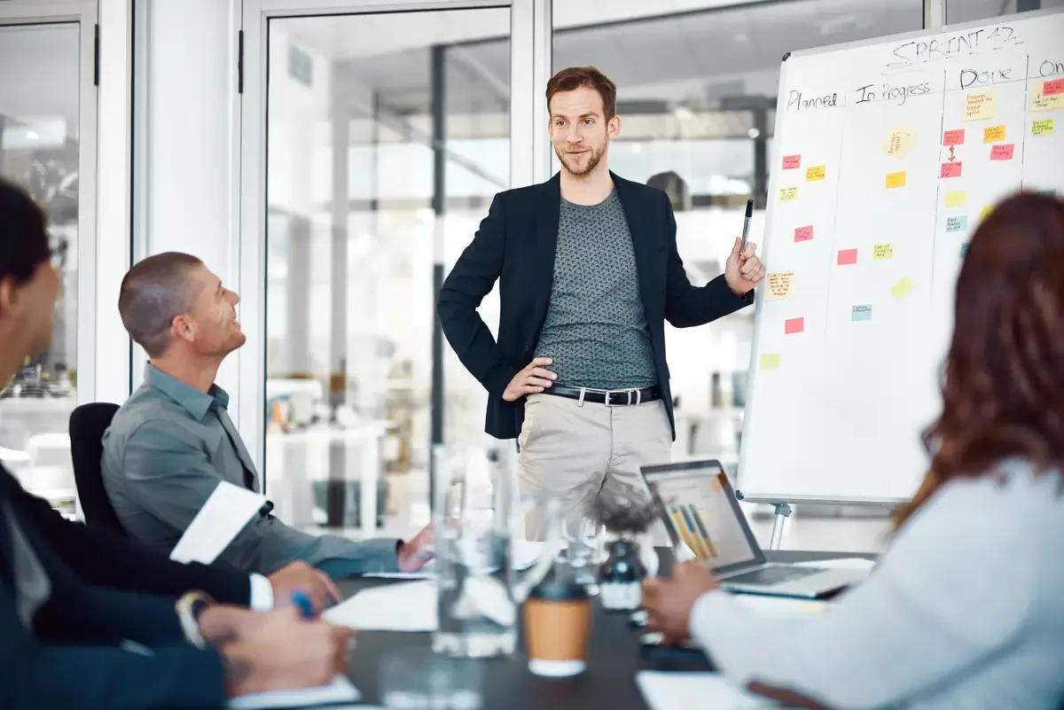A man presents a pitch deck to three attentive listeners in a modern glass-walled office.