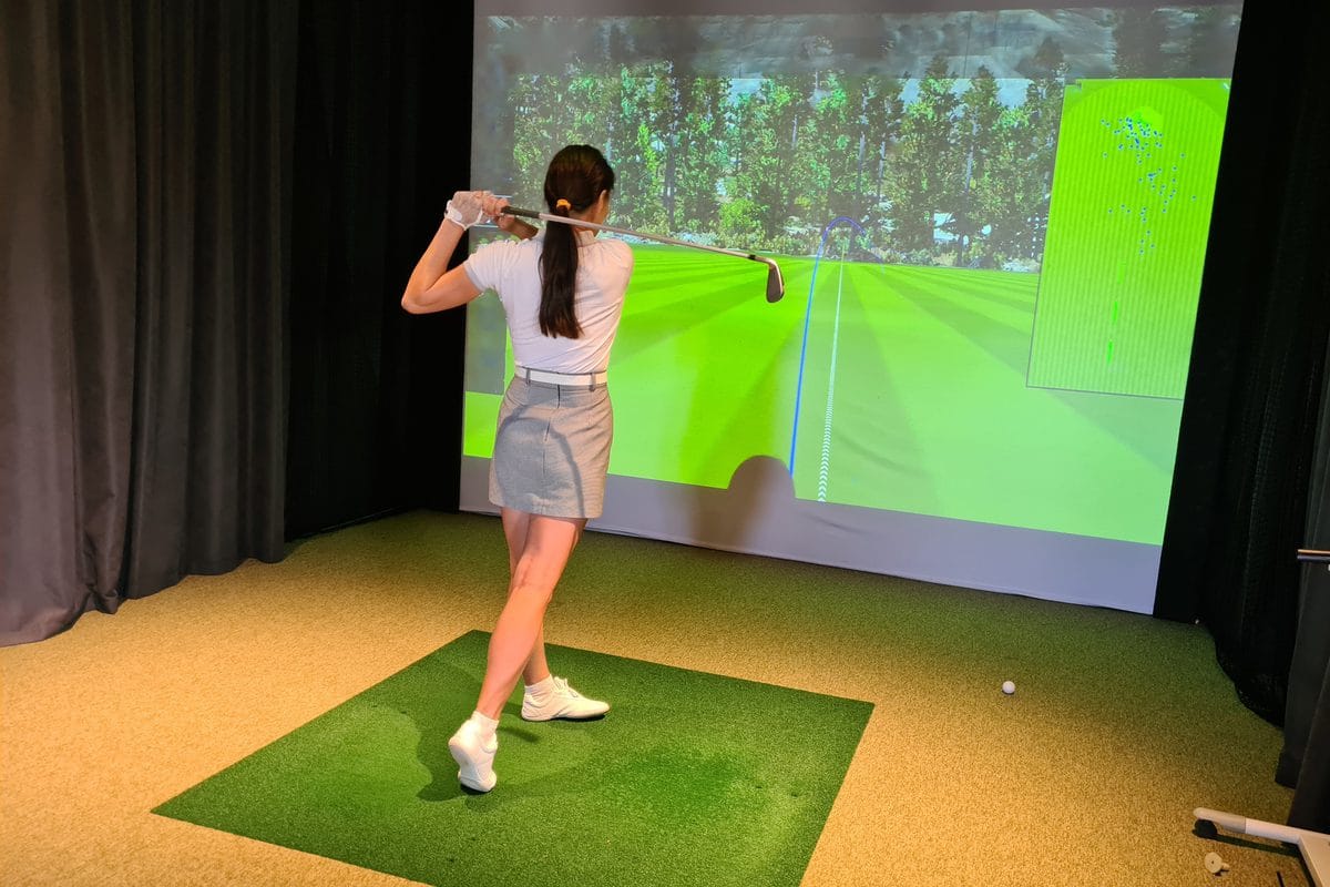 A golfer finishing a swing on a hitting mat in front of an impact screen