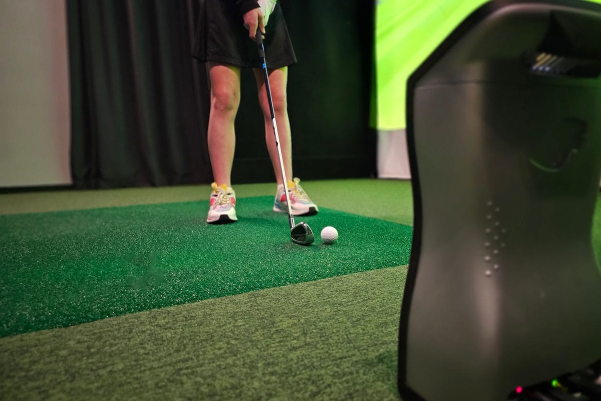 A floor-standing launch monitor beside a golf ball on a hitting mat in a simulator bay