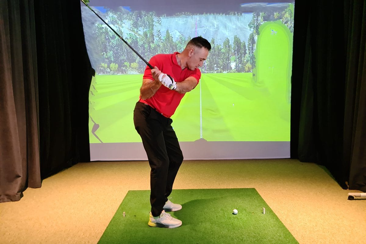 A golfer at the top of the backswing in an indoor simulator with dark side curtains