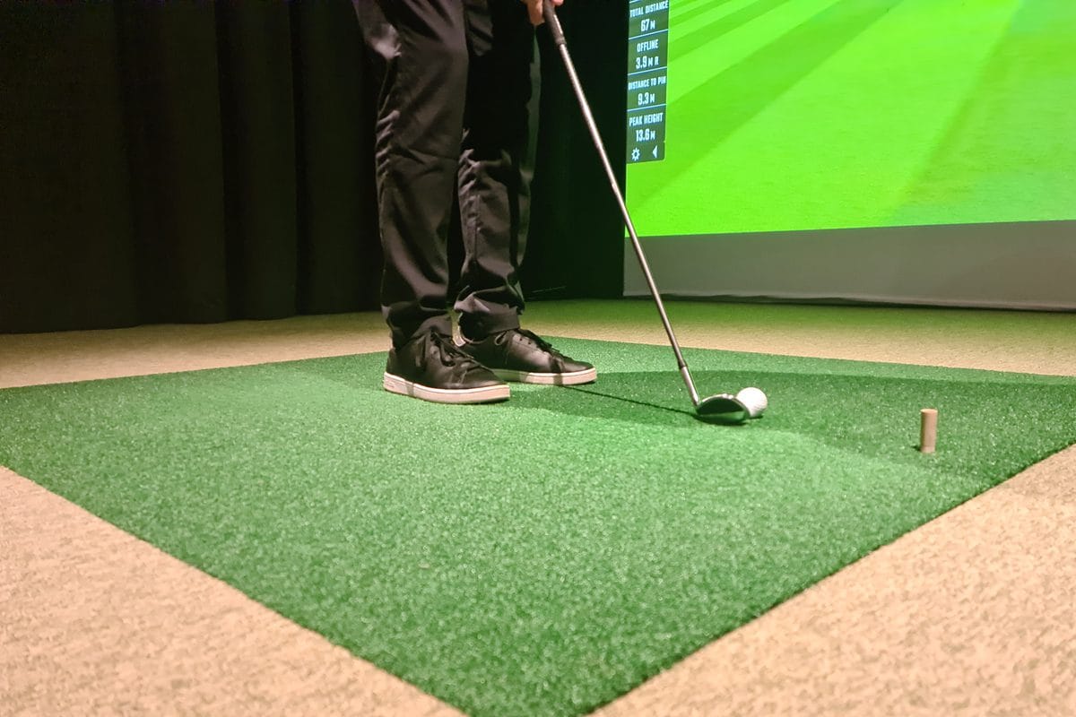 A golfer addressing a ball on a green hitting mat with the impact screen a few feet ahead