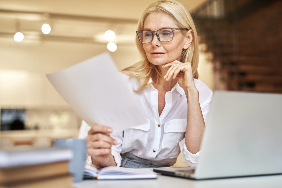 A woman at a desk reading a printed document beside a laptop