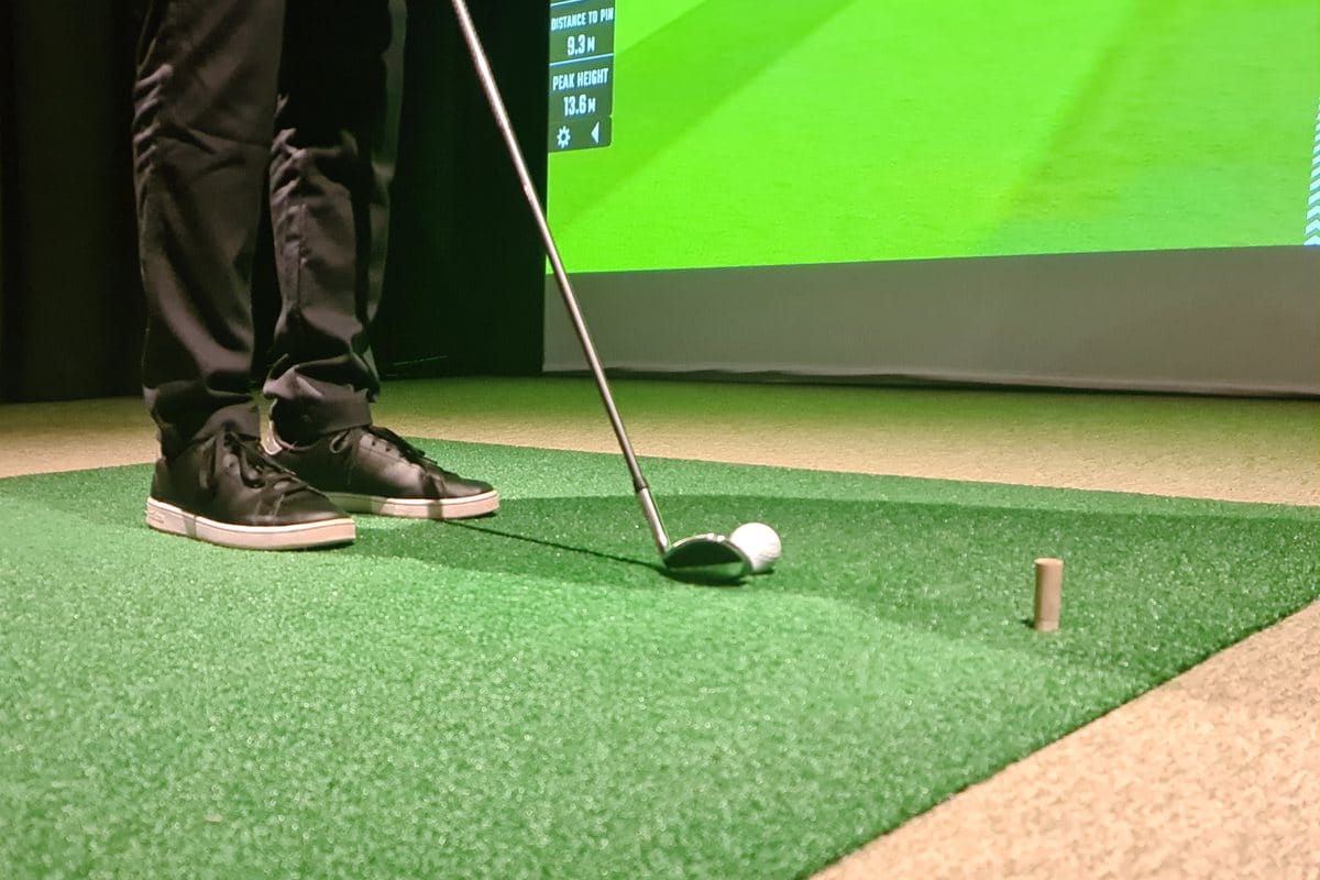 A golfer's shoes and an iron at address behind a ball on a turf hitting mat, a tee beside it, with a simulator screen behind