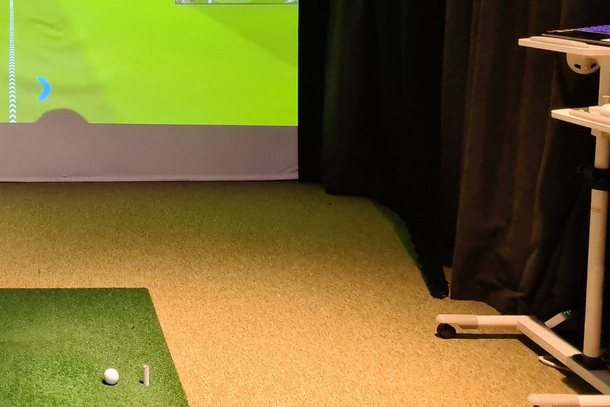 A rolling cart with a keyboard beside a golf simulator screen and hitting mat