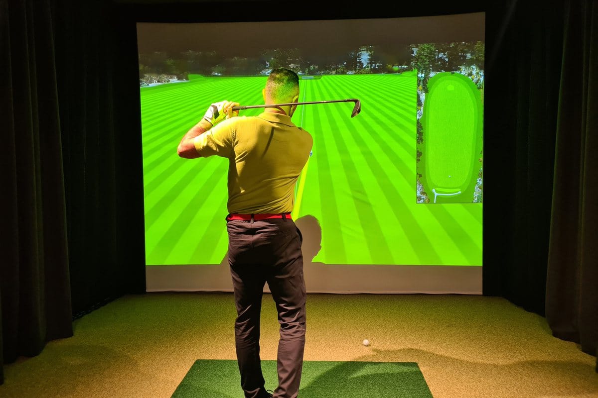 A golfer mid-swing in a home simulator bay with black curtains, an impact screen and a hitting mat