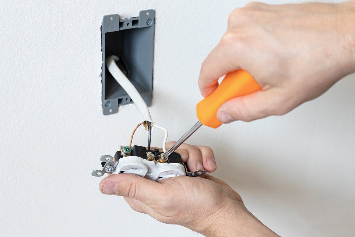 A hand with a screwdriver wiring a white duplex receptacle in front of an open wall box, the bare ground wire on its green screw