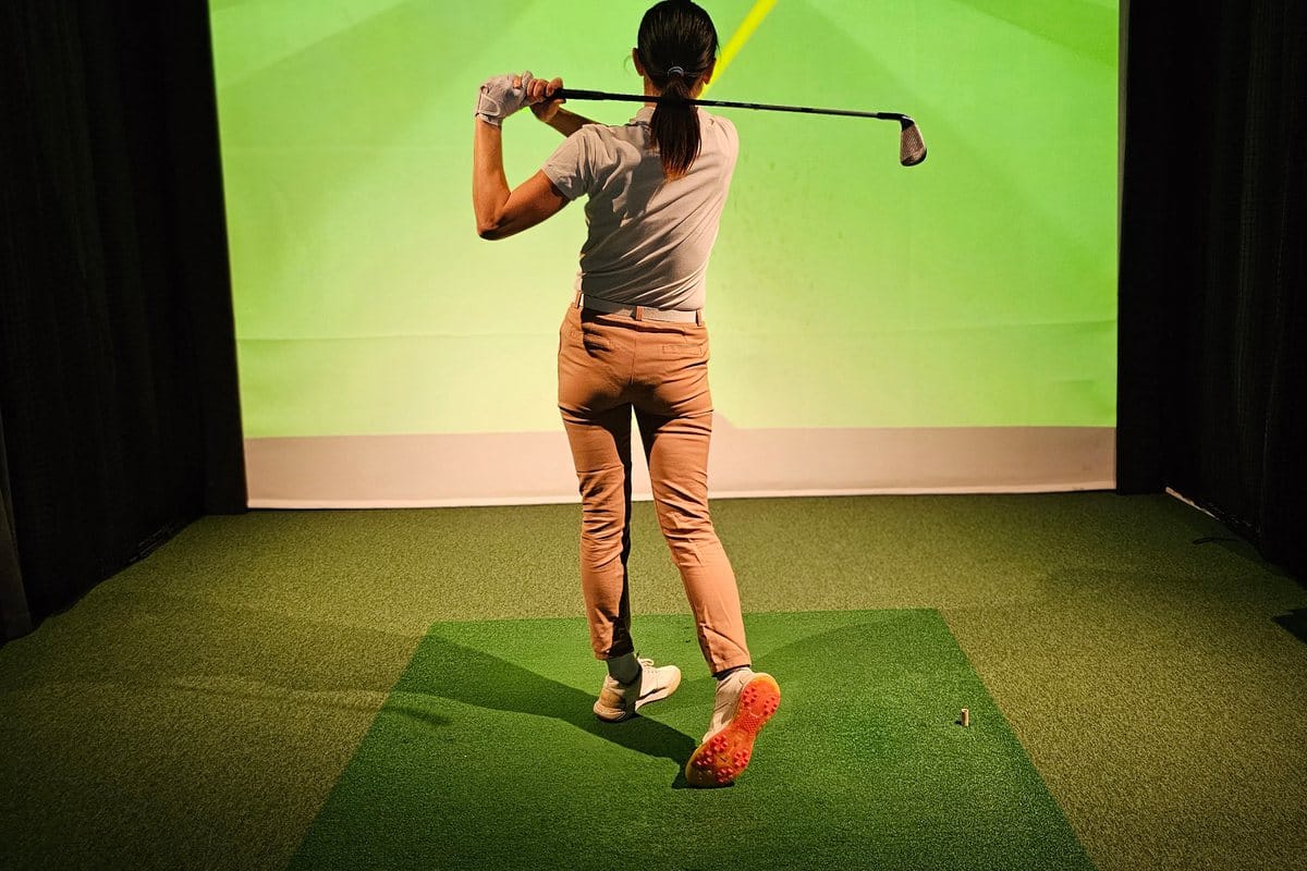 A golfer at the top of her follow-through on a turf mat, facing a lit simulator screen between black curtains