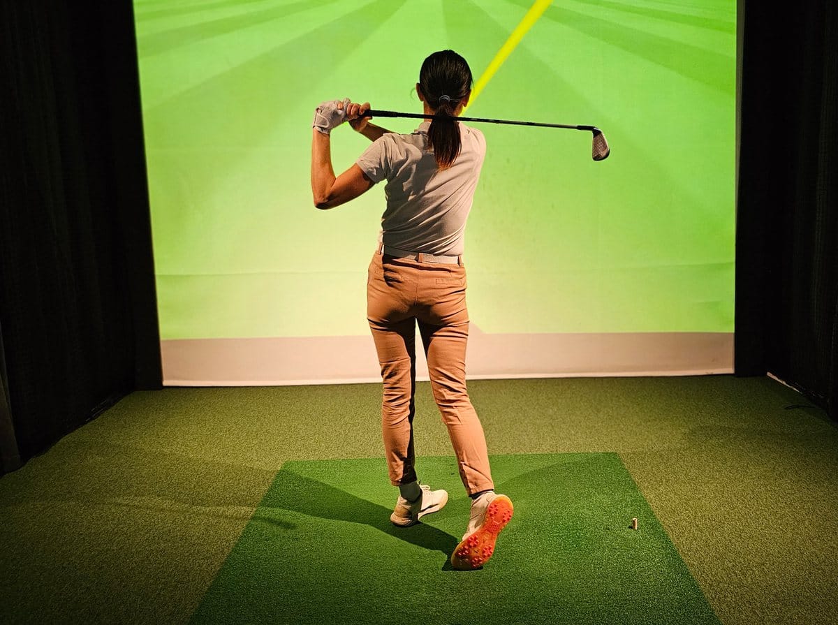 A golfer finishing a swing on a turf hitting mat in front of a lit simulator screen between black curtains