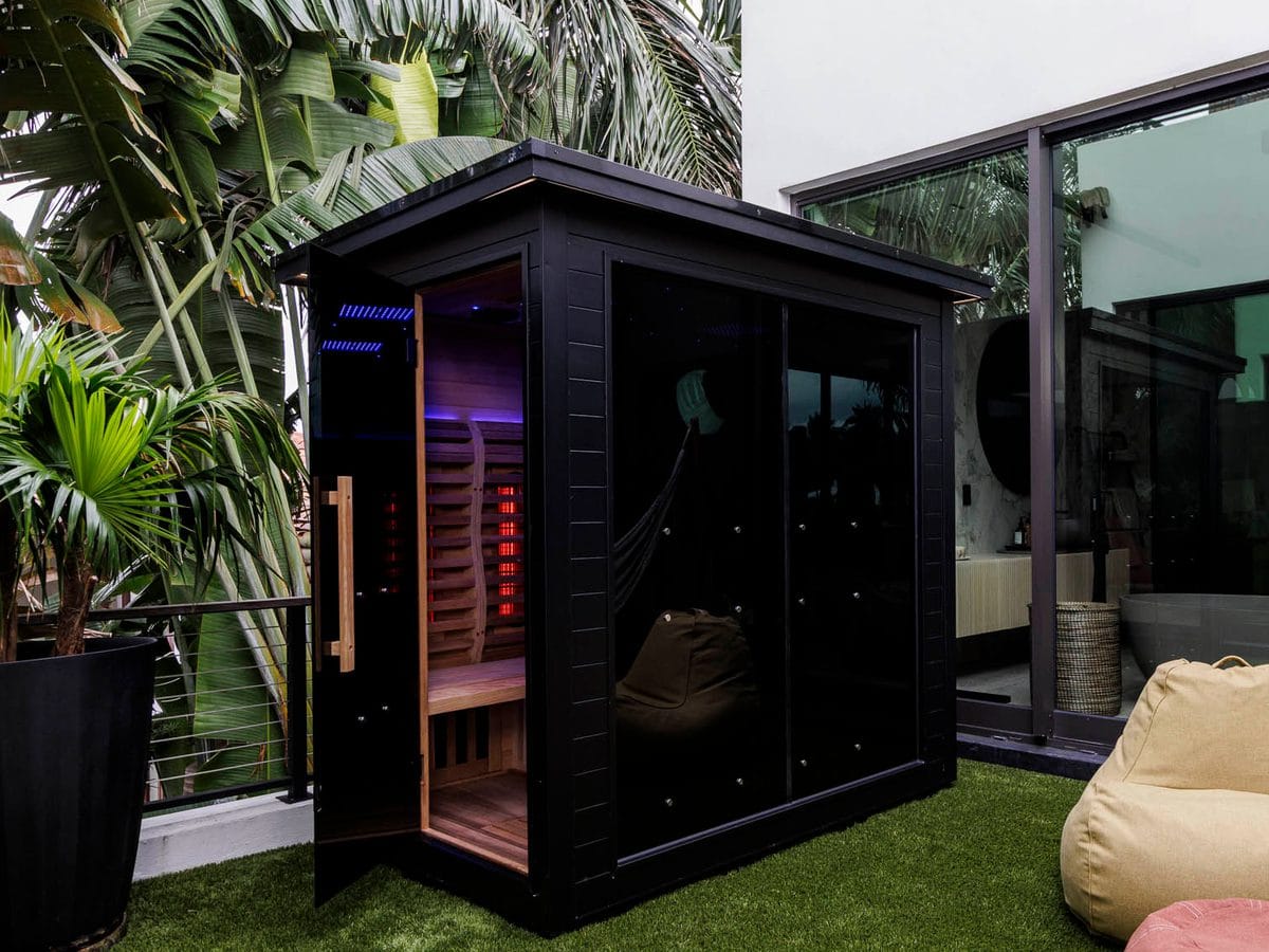 A black Sun Home Luminar 5 sauna with its door open to a wood bench and lit heater panels, on a backyard patio